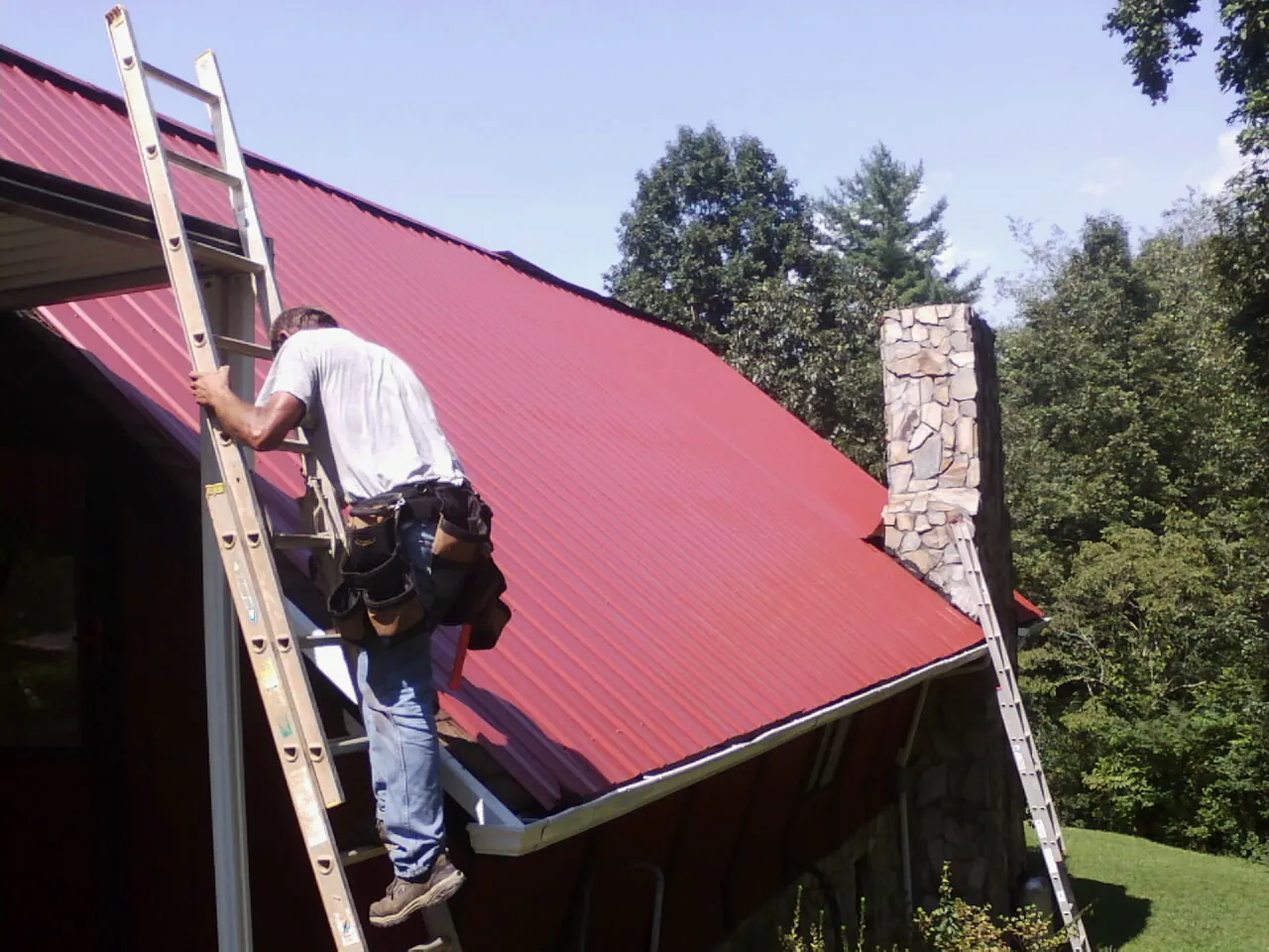 Roofer on a ladder beside a red metal roof and stone chimney — Soffit & Fascia Repair by Roofing Brothers in Scottville